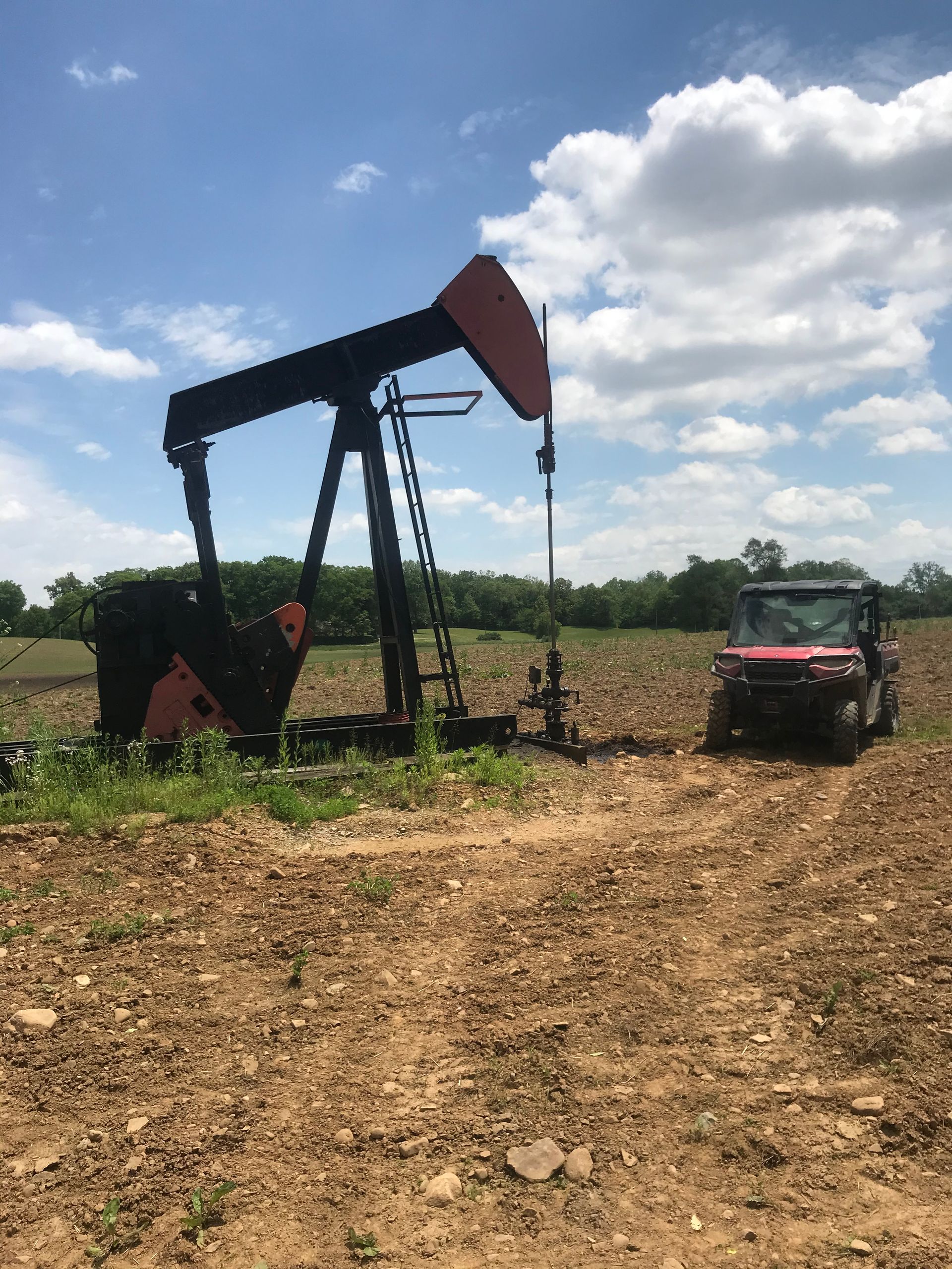 Oil pump jack in a field next to a red utility vehicle under a partly cloudy sky.