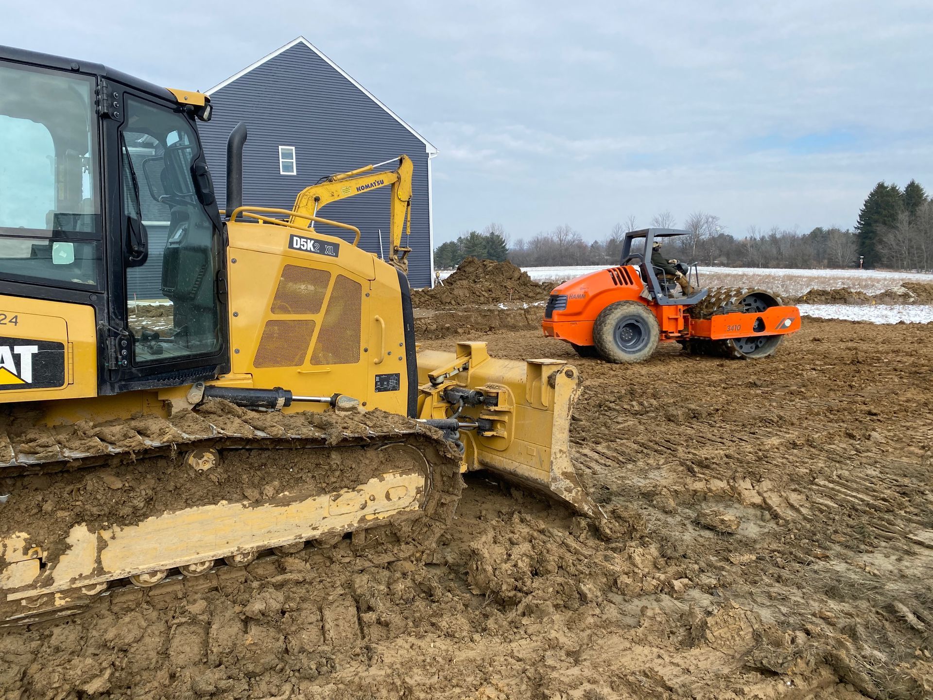 A yellow Caterpillar bulldozer and an orange roller compacting mud at a construction site.