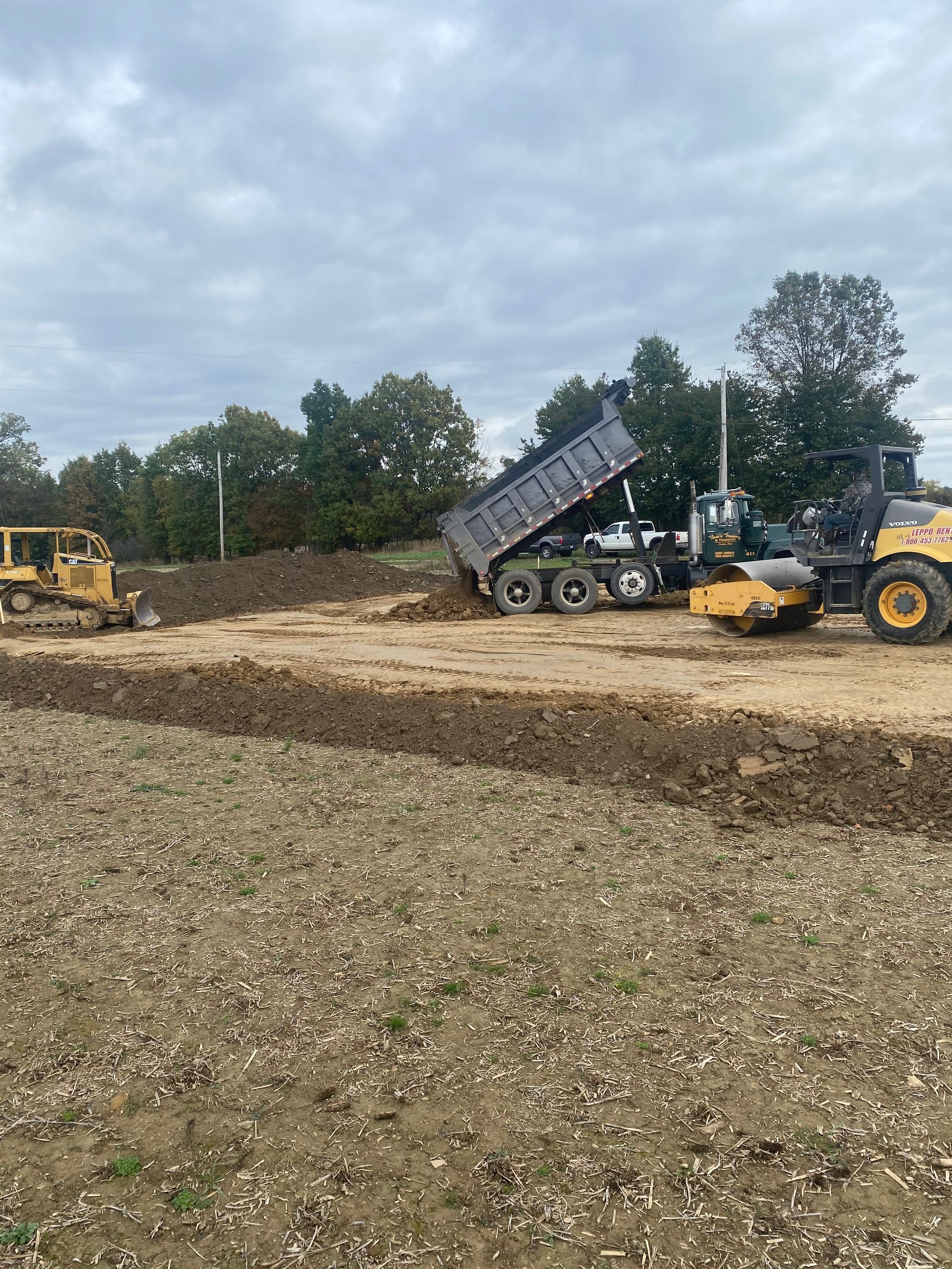 Dump truck unloading dirt onto a construction site with a grader in the background under an overcast sky.