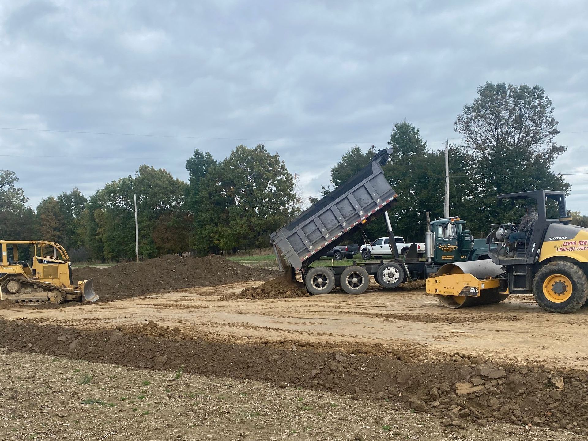 Construction site with a dump truck unloading dirt next to a compactor and bulldozer.