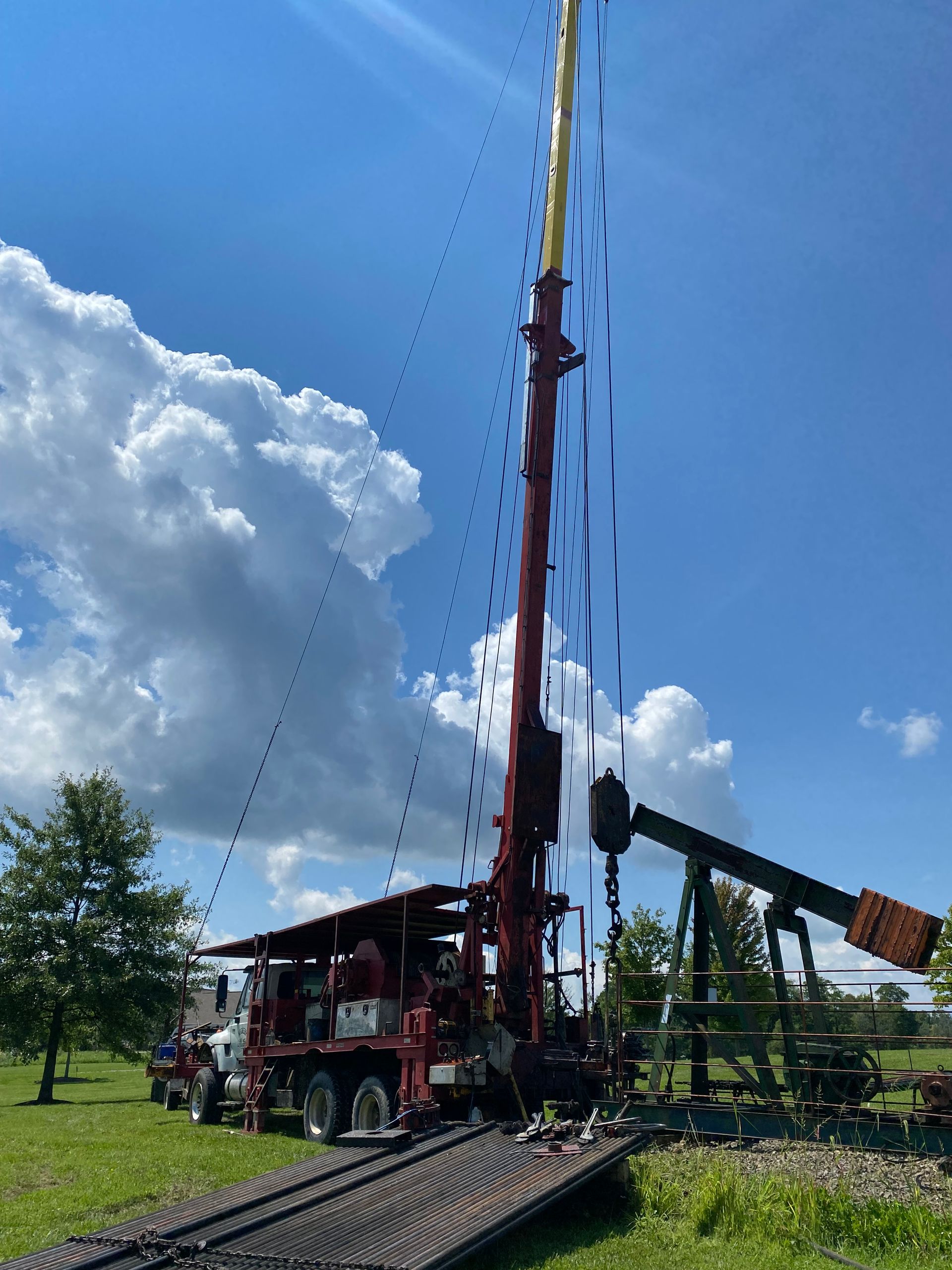 Drilling rig alongside an oil pump in a grassy field under a partly cloudy blue sky.