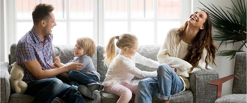 Family laughing together on a couch in a brightly lit room.