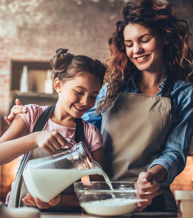 A person and a child wearing aprons smiling while pouring milk from a glass pitcher into a mixing bowl in a kitchen.