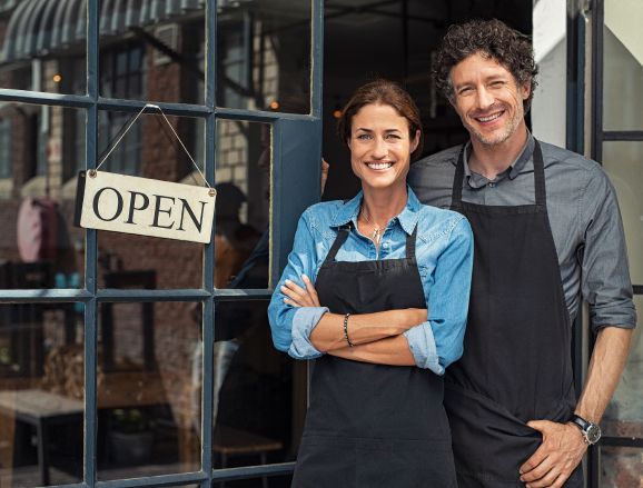 Two people wearing aprons smiling in front of a shop with an 