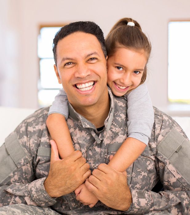 A smiling soldier in camouflage uniform being hugged from behind by a child.