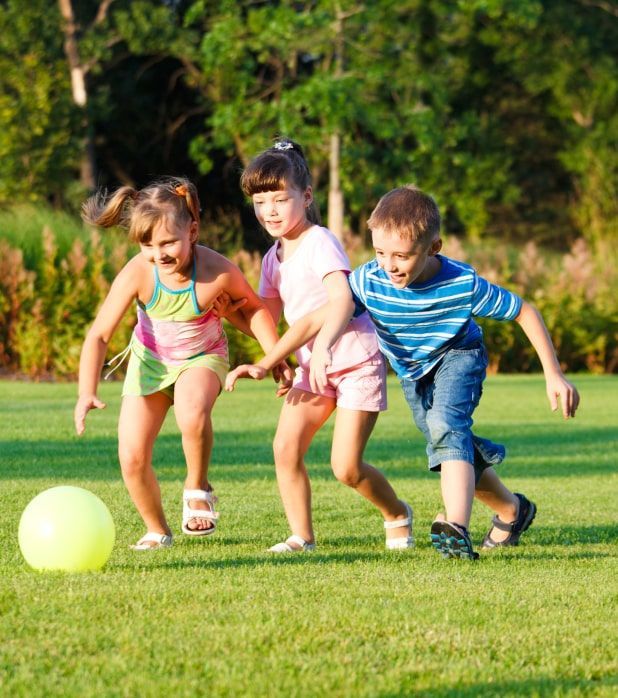 Three children running across a sunlit grass field while chasing a large yellow ball.