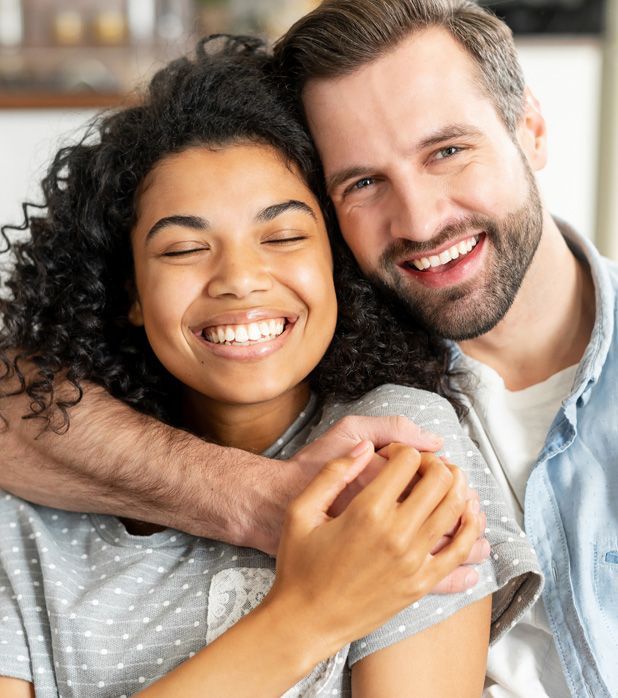 A smiling person with dark curly hair is embraced by another person from behind, both looking happy in a close-up shot.