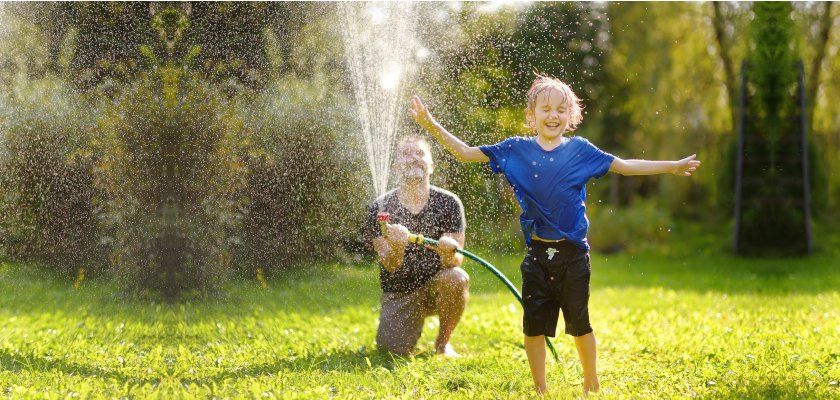A person kneels, spraying water on a smiling child with arms outstretched on a grassy lawn.