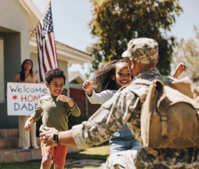 Soldier in uniform hugs two children running towards him, wife holding welcome sign, American flag in background.