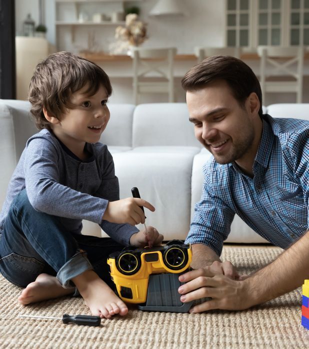 A parent and child sit on a living room rug, smiling as they use a small screwdriver to repair a yellow toy vehicle.