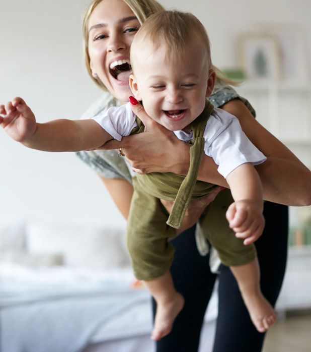 Woman holding a laughing baby wearing green overalls and a white shirt.