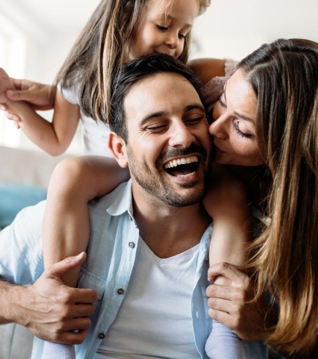 A laughing man gives a child a piggyback ride while a woman kisses his cheek.