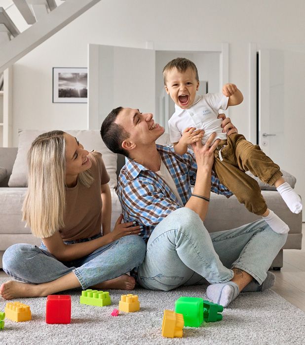 A family sits on a living room rug, playing with colorful building blocks as a parent lifts a laughing toddler.