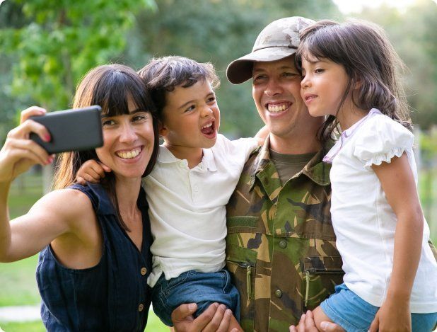 Family taking a selfie; father in camouflage, mother holding phone, children smiling outdoors.