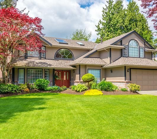 Tan two-story house with red door, large green lawn, and colorful landscaping.