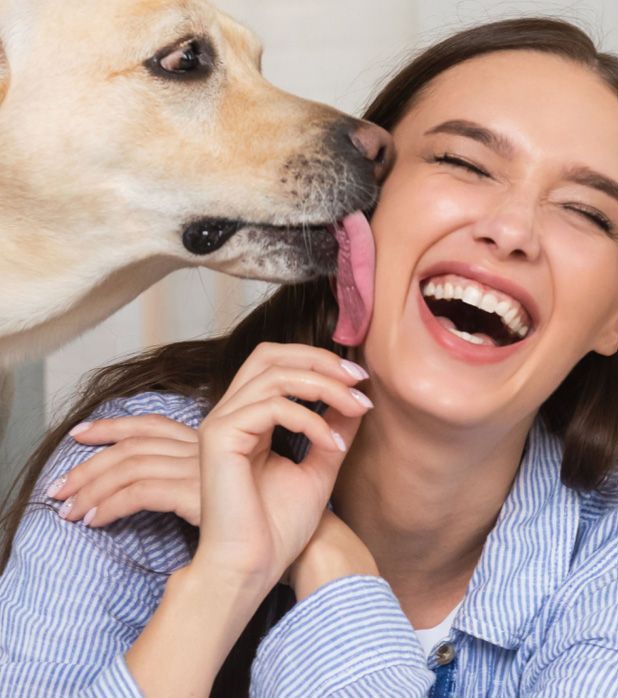 A light-colored dog licks the cheek of a joyful person who is laughing with their eyes closed.