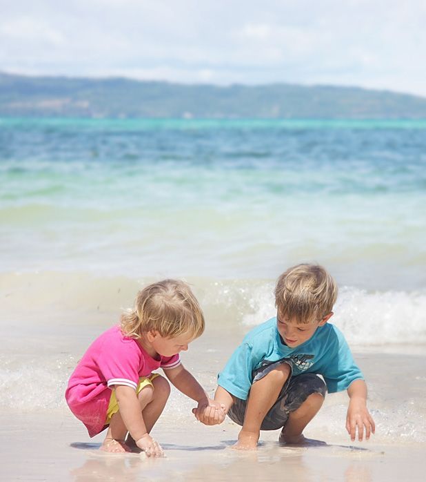 Two children holding hands while crouching on a sandy beach by the ocean.