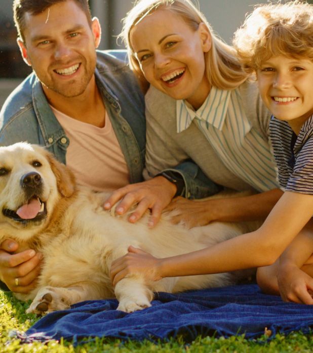 A family and their golden retriever smile while sitting together on a blue blanket on a sunny lawn.