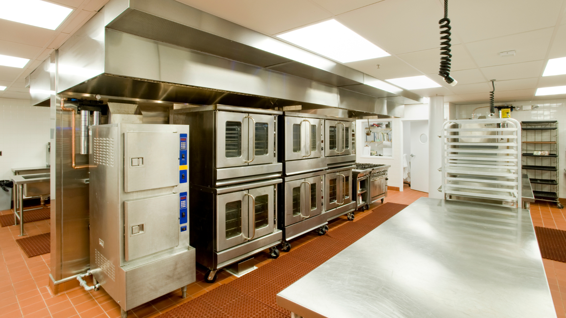 Commercial kitchen with stainless steel appliances, ovens, and vent hood. Red tile floor.