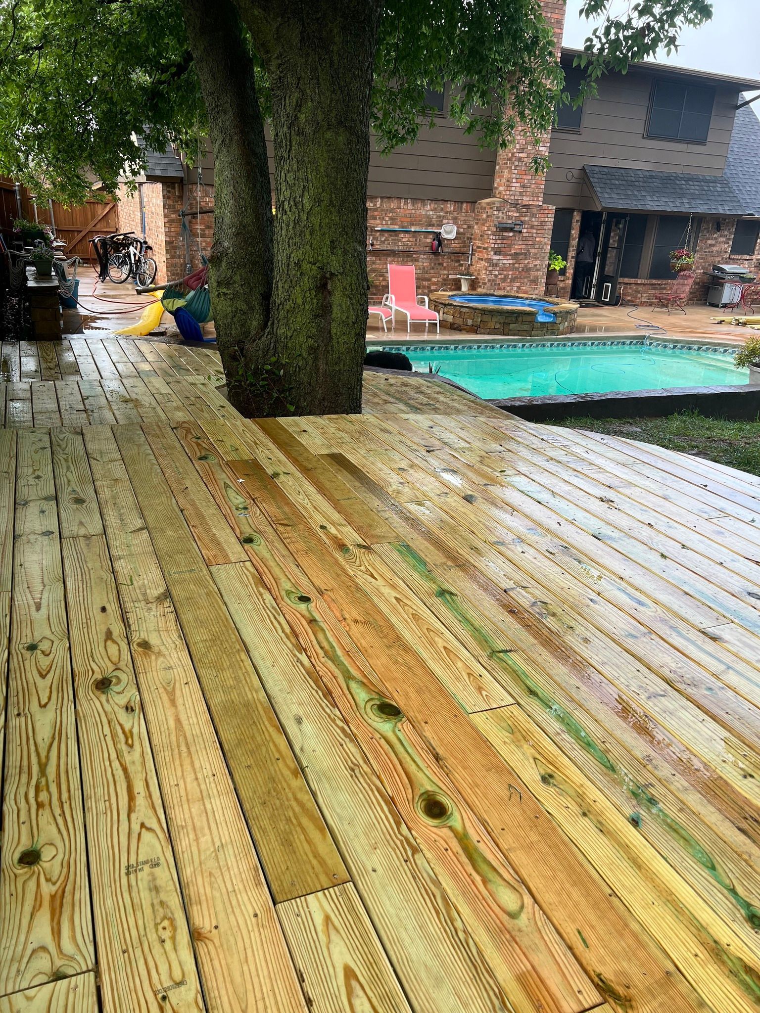 Wooden deck around a tree near a pool, with a brick house in the background.