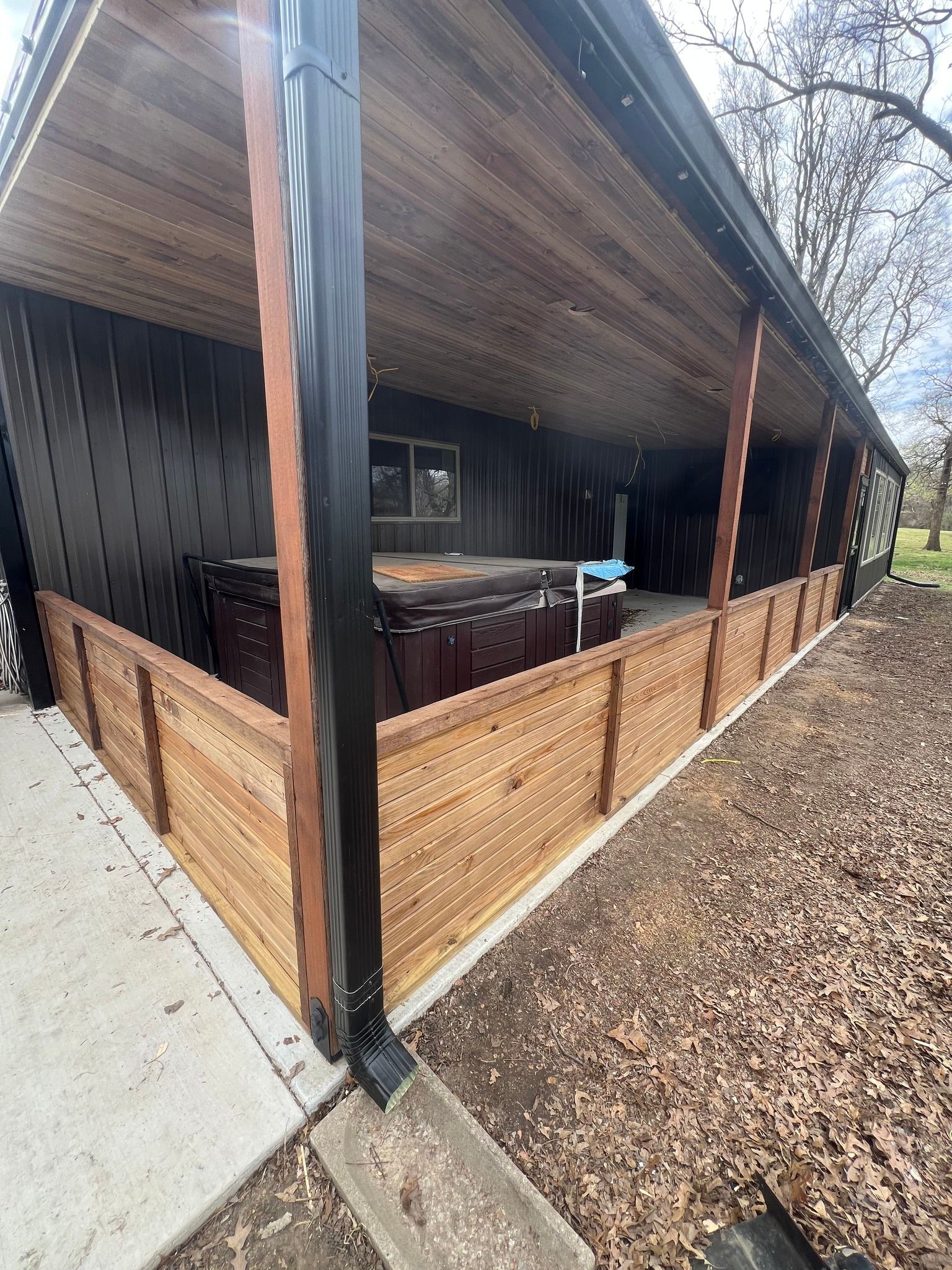 A covered outdoor hot tub area with a wooden fence and dark siding under a roof.