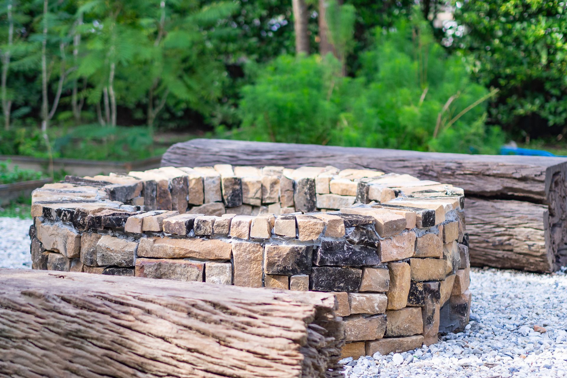Circular stone fire pit with wooden logs, gravel ground, and greenery in the background.
