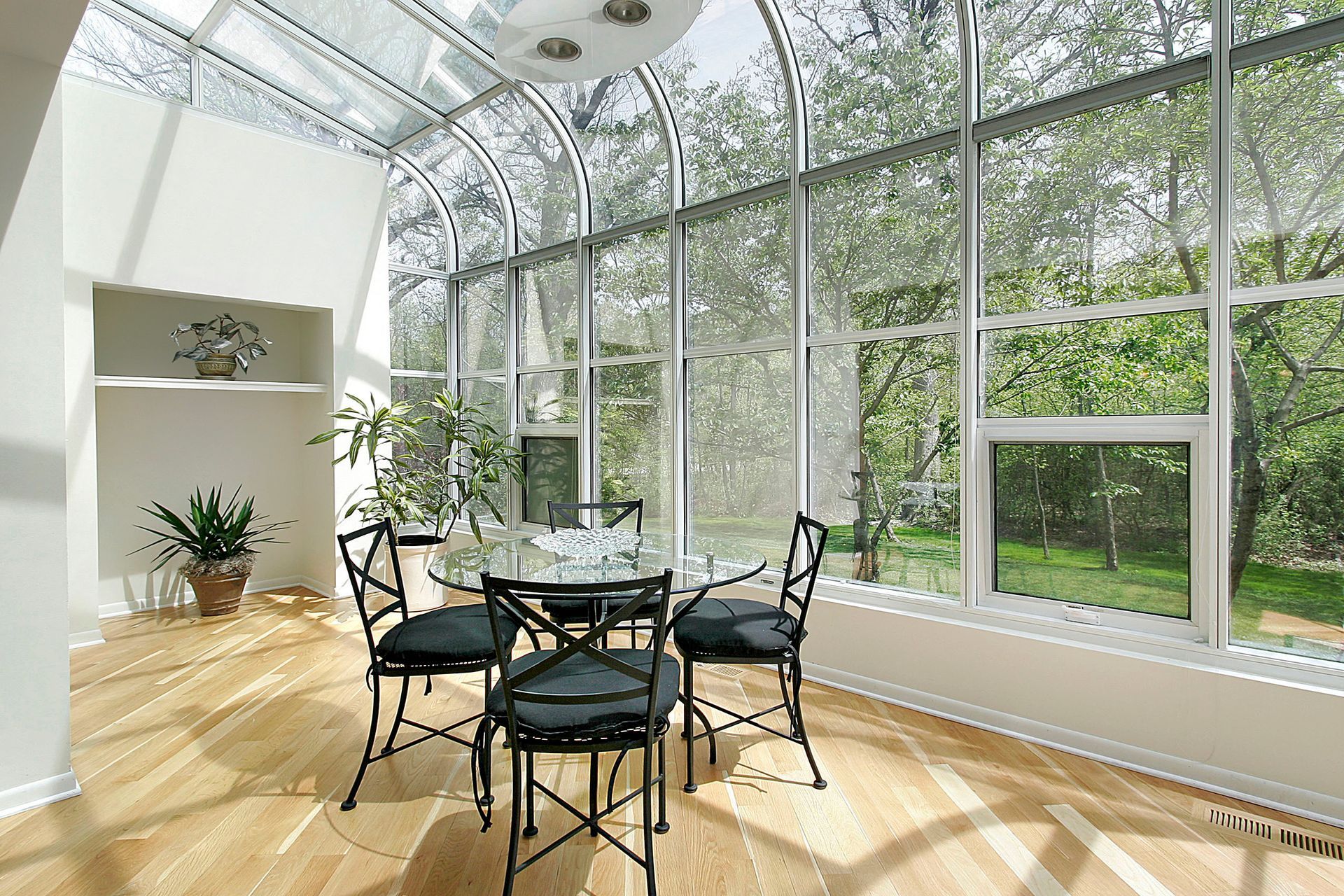 Sunroom with glass ceiling and walls, round table with chairs, hardwood floor, and plants.
