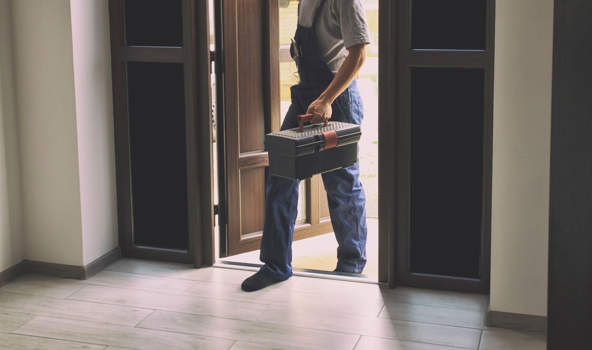 Person in blue overalls entering a building, carrying a toolbox through an open doorway.