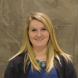 Woman with long blonde hair smiles, wearing blue top and necklace, standing against a patterned wall.