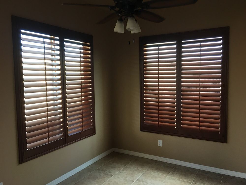 Two windows with wood shutters in a tan-colored room; ceiling fan in the corner; beige floor.
