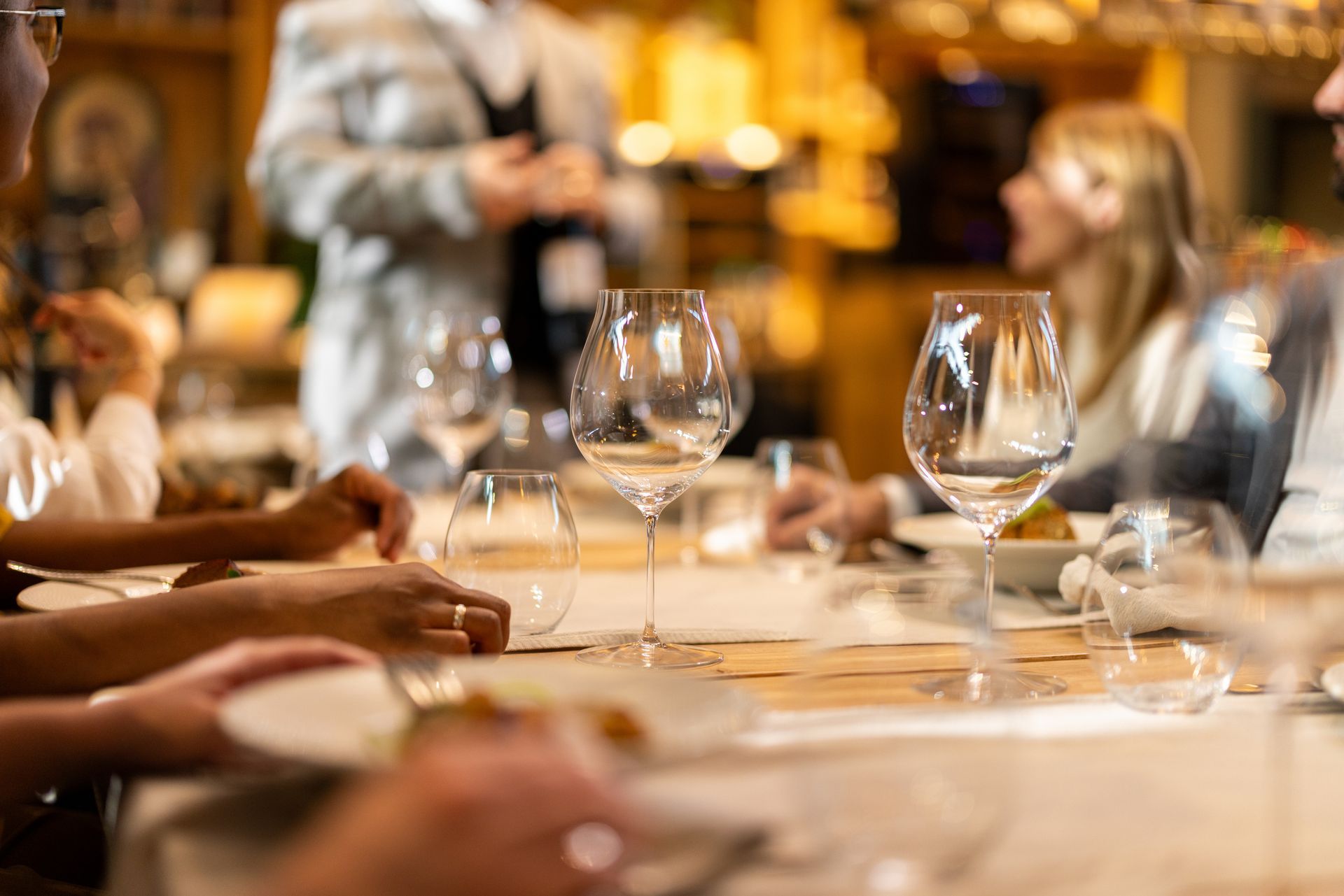 Tavolo da pranzo apparecchiato con bicchieri da vino, persone sedute, cameriere che versa il vino nel ristorante.