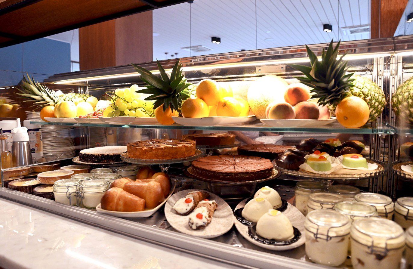 Esposizione di dolci con torte, frutta e piccoli barattoli sotto luci intense.