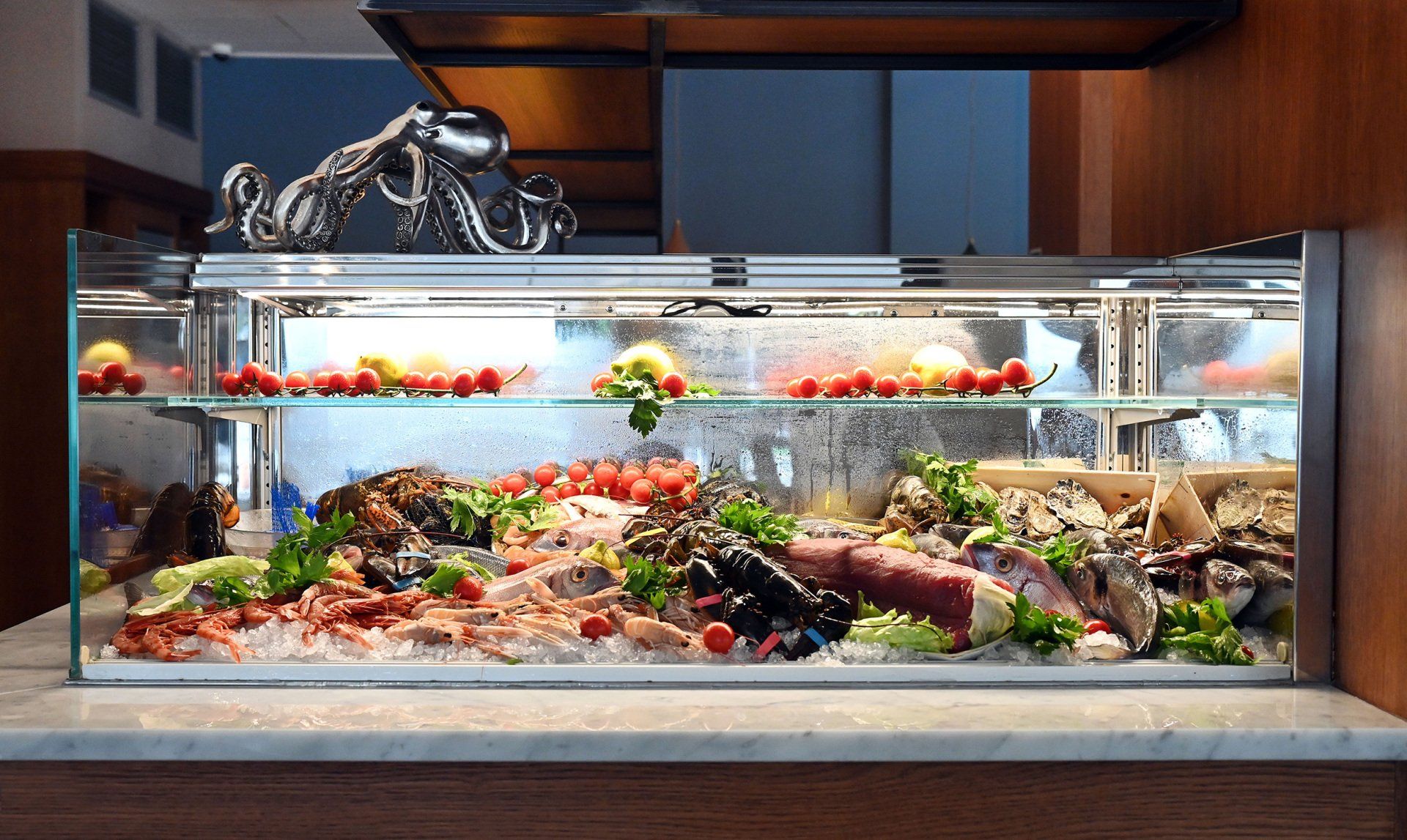 Vetrina per frutti di mare su un bancone in marmo con una scultura di polpo sopra, piena di pesce fresco e prodotti agricoli.