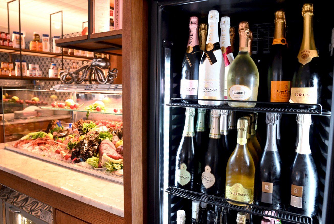 Vetrina refrigerata con bottiglie di spumante, accanto a un buffet di cibo fresco.
