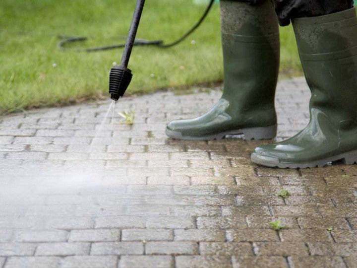 A person wearing green boots is using a high pressure washer to clean a brick walkway.