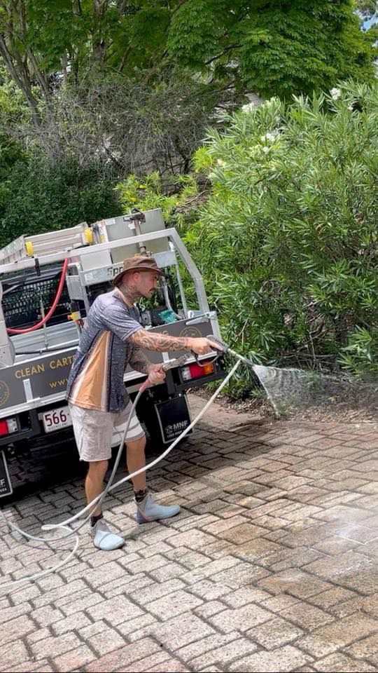 A Man Is Spraying Water On A Brick Driveway With A Hose — Clean Cartel Pressure Washing In Reedy Creek, QLD