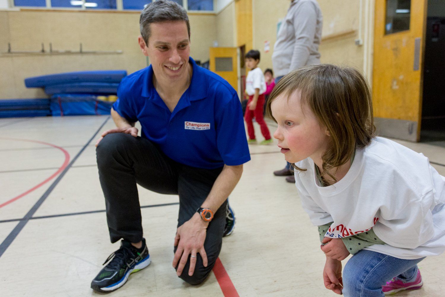 A man is squatting down next to a little girl in a gym.