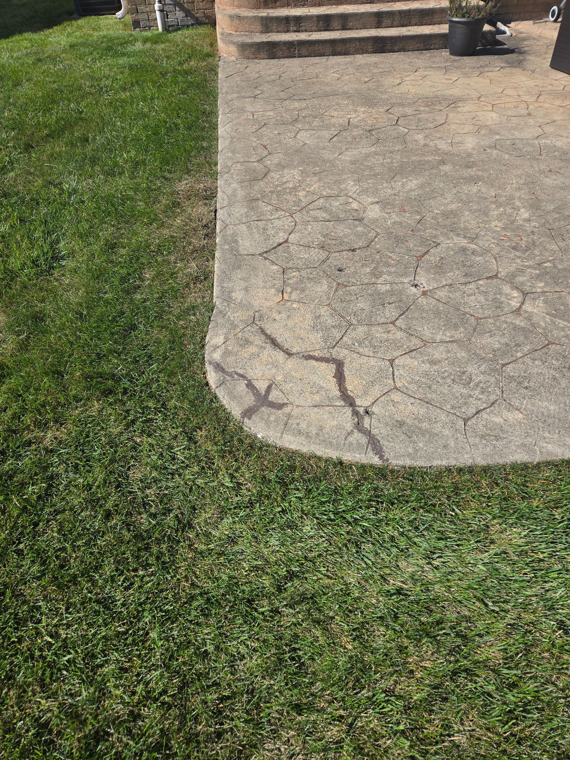 Concrete patio corner with visible cracks and surface wear, set against a patch of green grass.