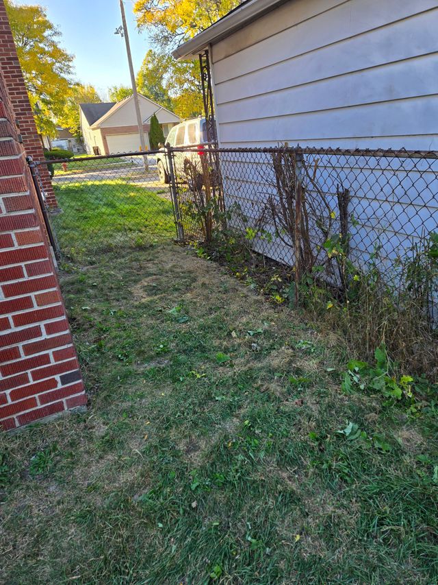An outdoor view of a narrow grassy strip between a red brick wall and a chain-link fence, with a house visible behind it.