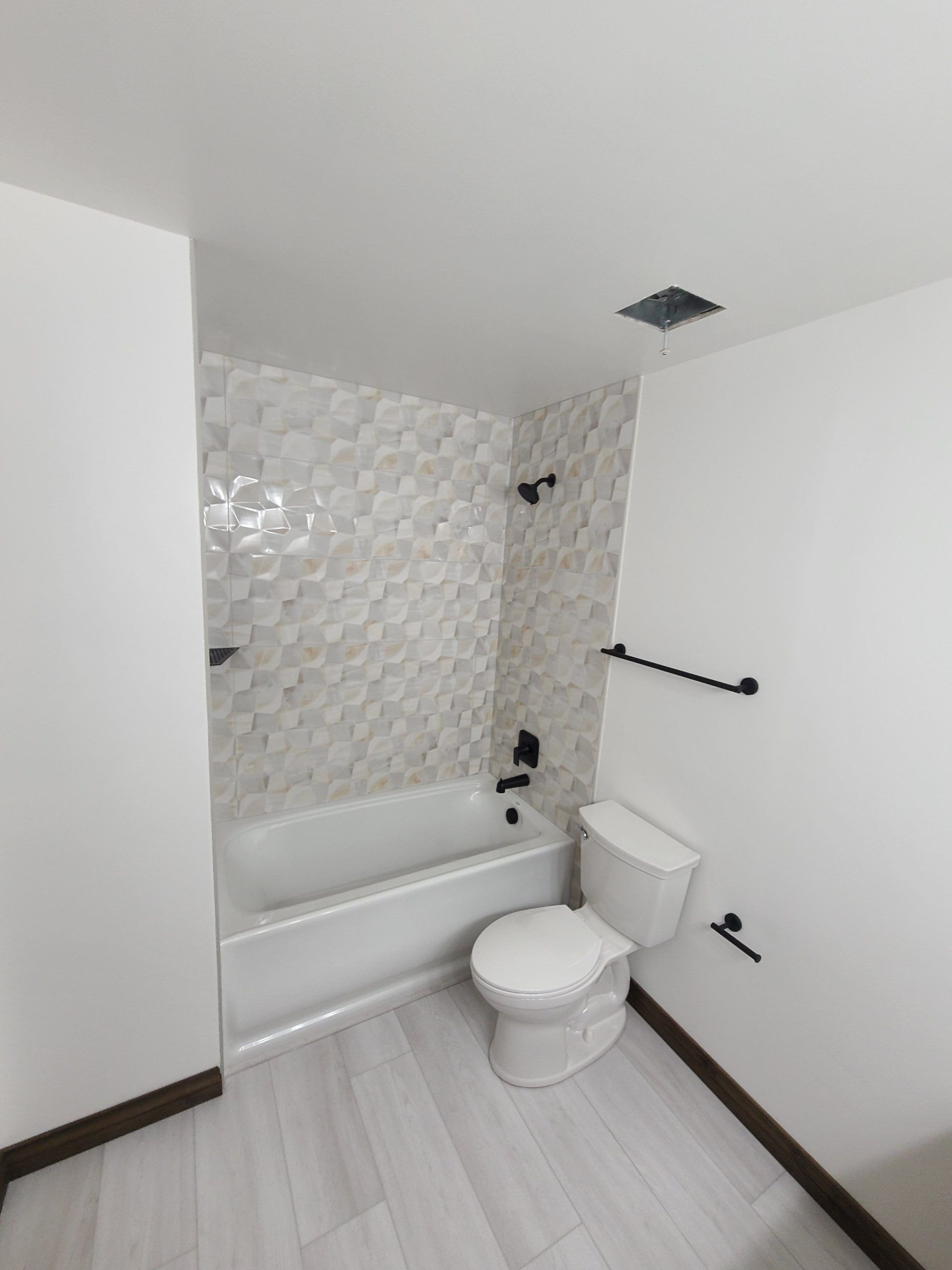 A white bathroom with a tub, toilet, and patterned tile wall. Black fixtures and light wood flooring.