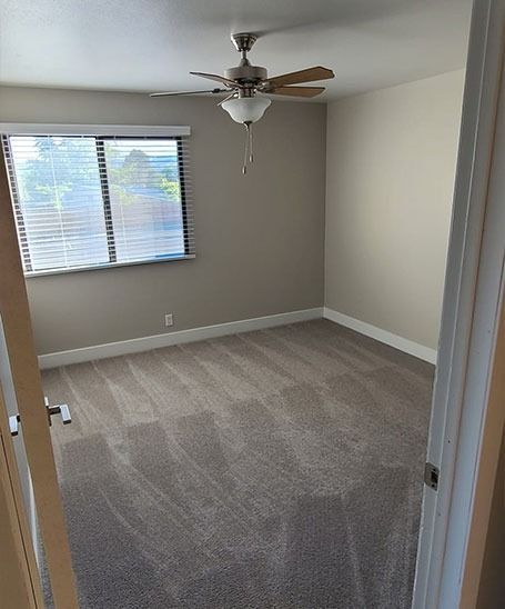 Empty bedroom with gray carpet, beige walls, blinds, and ceiling fan.