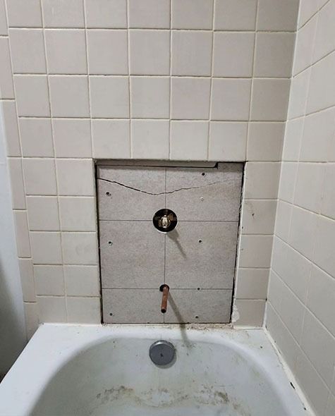 Bathtub with a square opening in the tiled wall. Exposed pipes and cracked tile are visible within the opening.