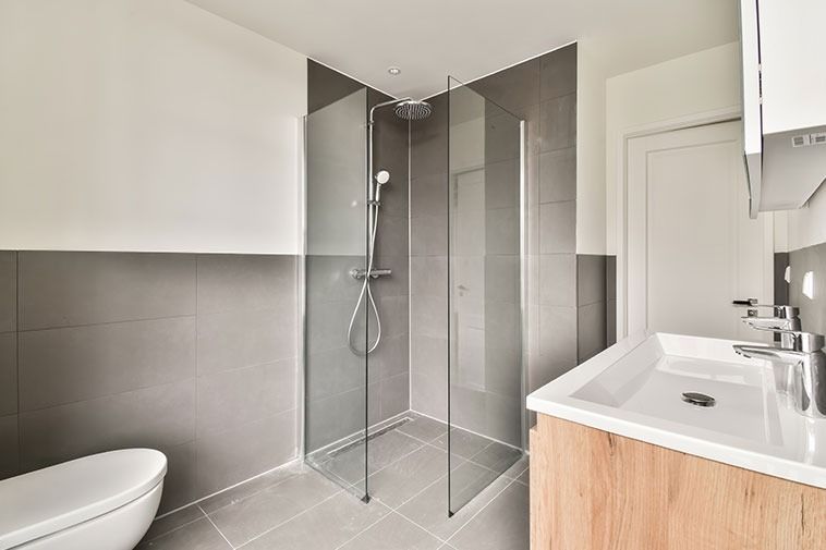 Modern bathroom with corner shower, gray tile, white toilet, and a light wood vanity.