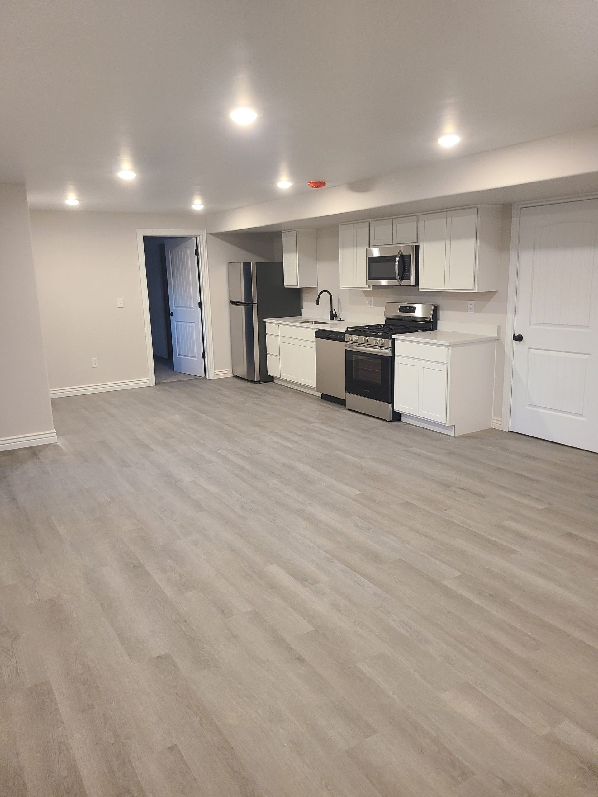 Empty, modern, light-floored kitchen and living area. Stainless steel appliances. White cabinets. Recessed lighting.