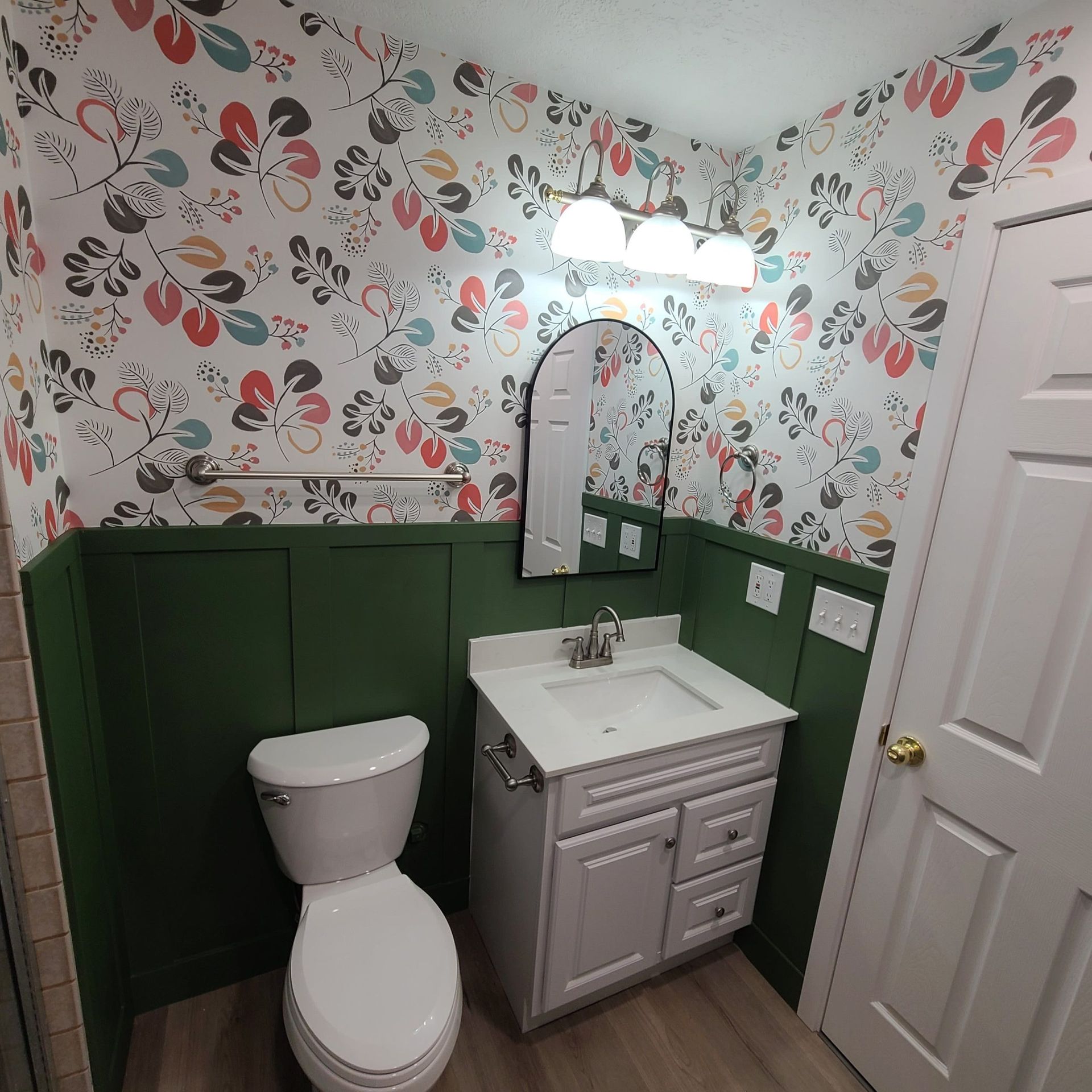 Bathroom with floral wallpaper, green wainscoting, white vanity, toilet, and a white door.