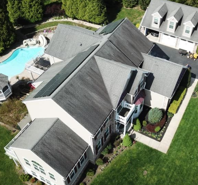 Aerial view of a large suburban house with a gray shingled roof experiencing dark streaking from water retention.
