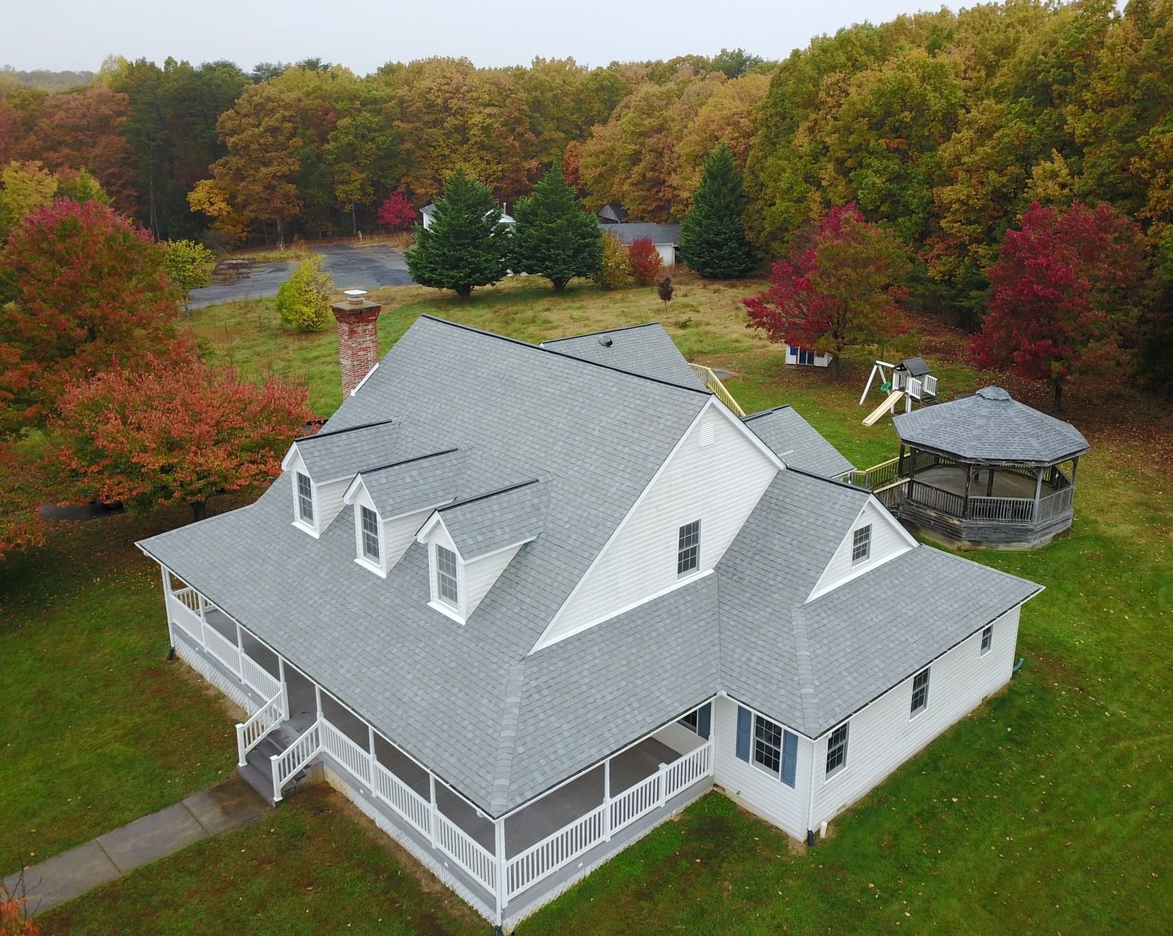 Aerial view of a gray-roofed house with a porch, surrounded by trees with fall foliage and a gazebo. Roof replacement by Four Seasons