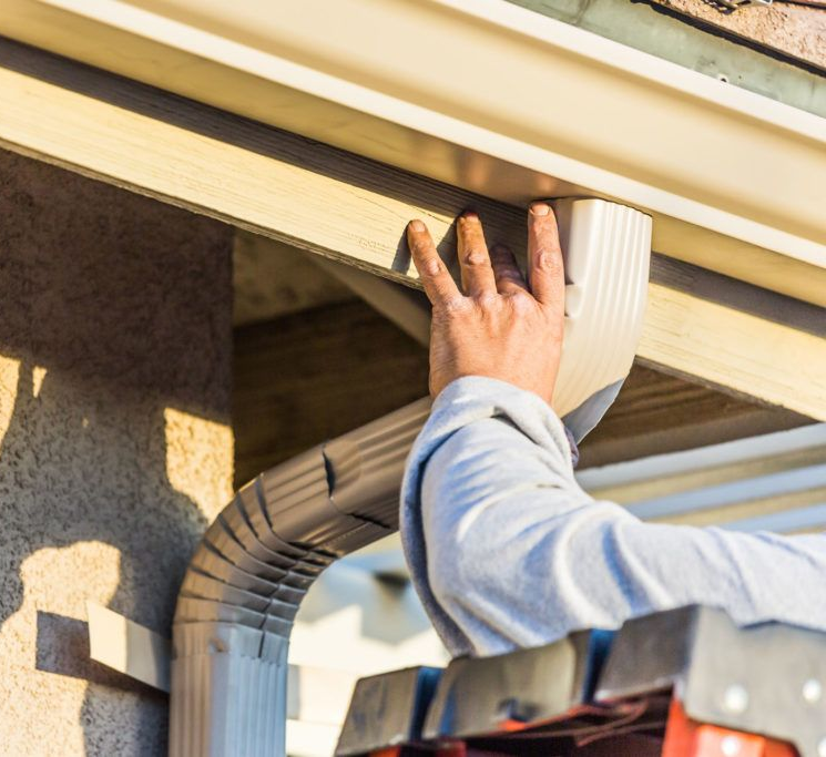 professional roofing contractor on ladder installing a beige gutter downspout.