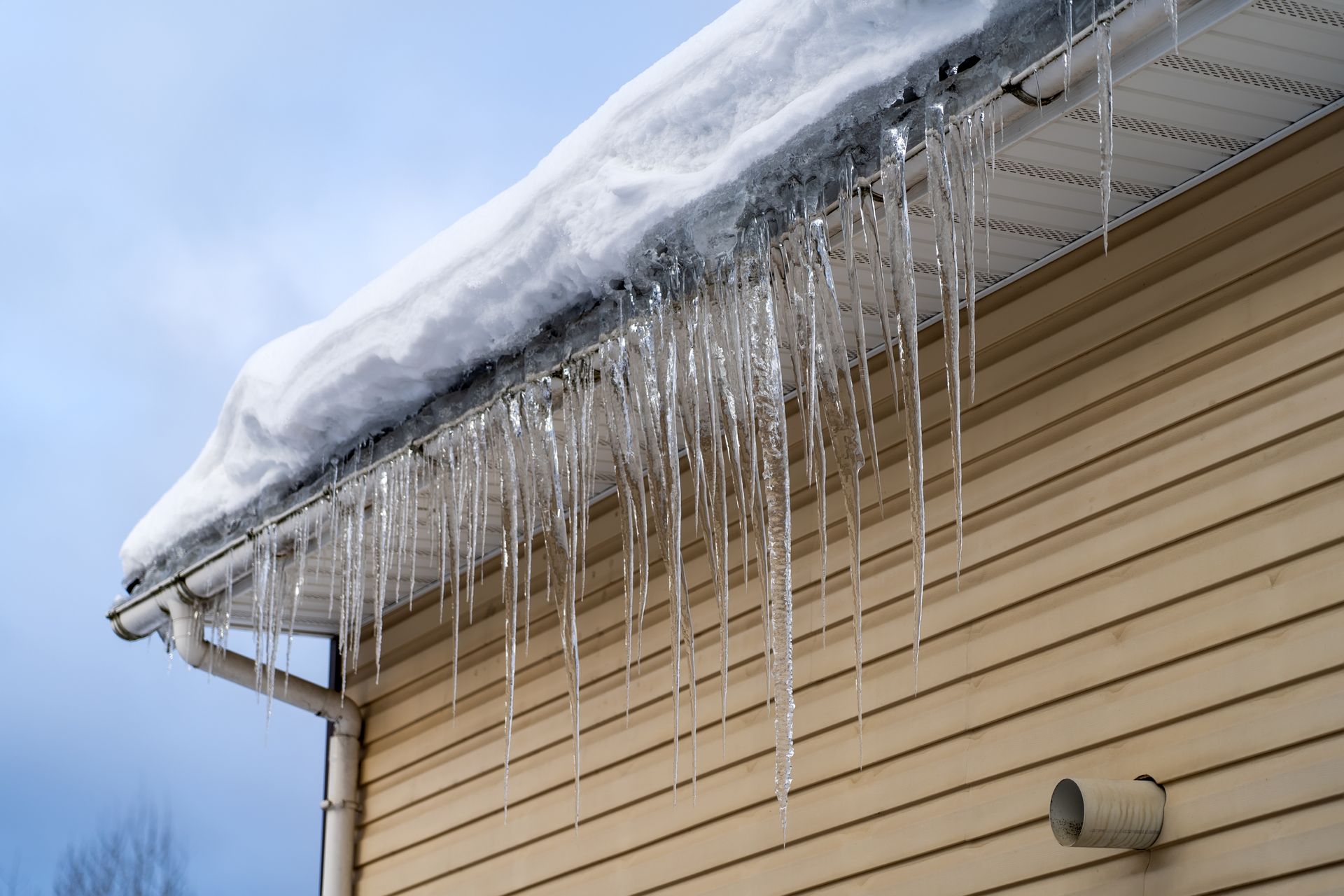 Snow-covered roof with ice dam, icicles hanging from the edge of the roof, beige siding below.
