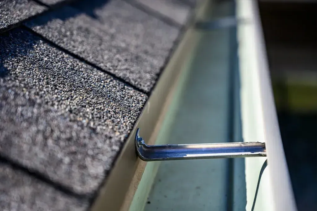 A metal hanger bracket secures a white rain gutter to the edge of a dark shingled roof.